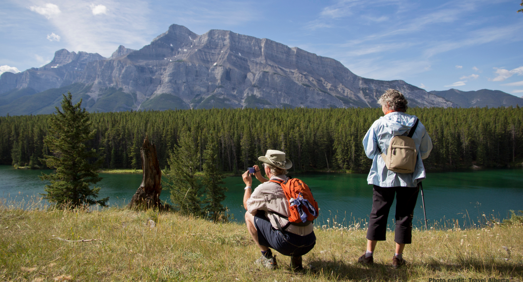 On a river bank is a couple taking photos of Banff National Park mountains.
