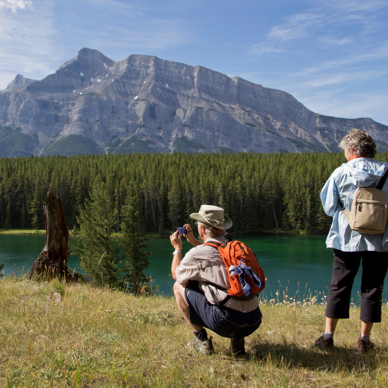 On a river bank is a couple taking photos of Banff National Park mountains.