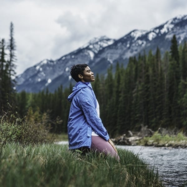 Girl sitting on her knees on the edge of river with the mountains in the background.