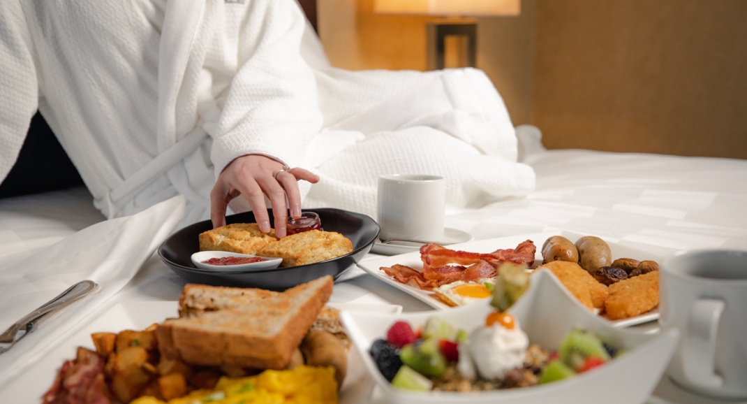 Women in a robe with a breakfast spread on the bed at The Rimrock Resort Hotel.