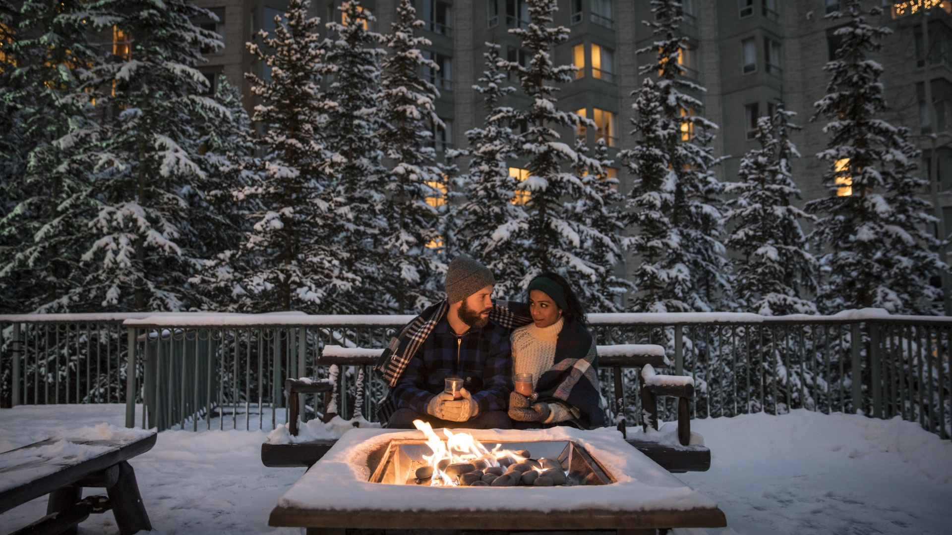 Couple sitting by the outdoor fire pit with snow covered trees in the background at The Rimrock Resort Hotel in Banff National Park