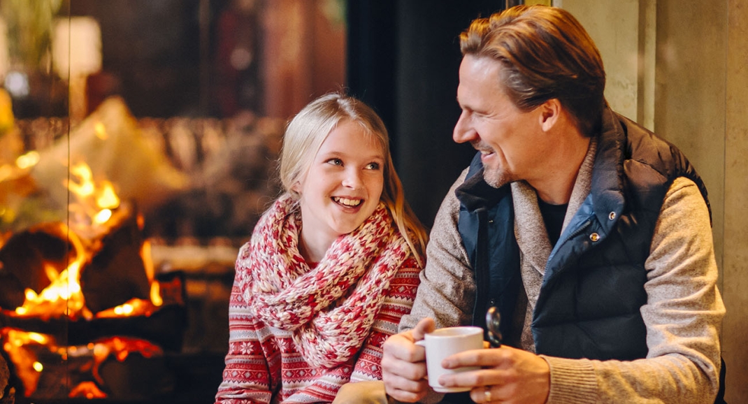 Dad and daughter sit by the fire around the festive season drinking cocoa at The Rimrock Resort Hotel.