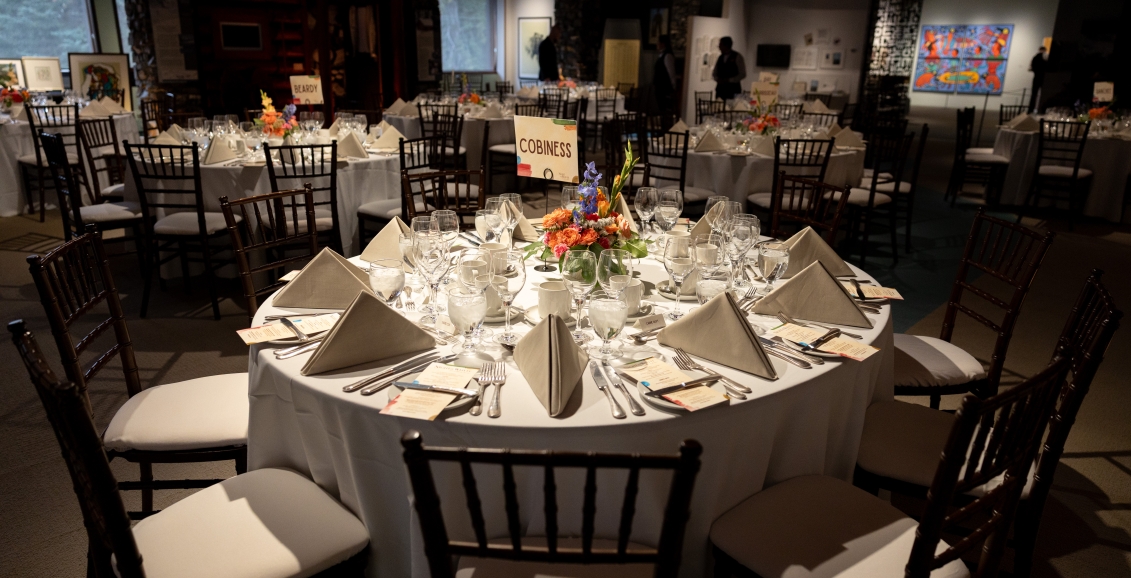Round banquet tables set for an elegant dinner at The Whyte Museum, featuring folded napkins, glassware, floral centerpieces, and wooden chairs in a softly lit gallery space.