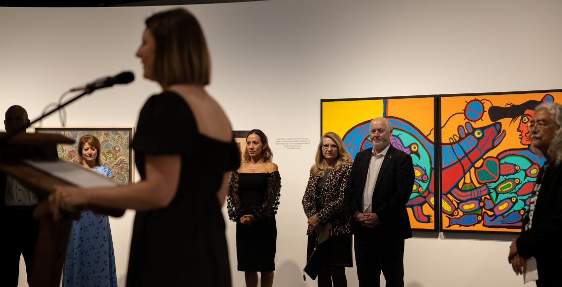 A speaker at a podium addressing attendees in an art gallery at The Whyte Museum, with guests standing in front of vibrant Indigenous artwork on the walls.