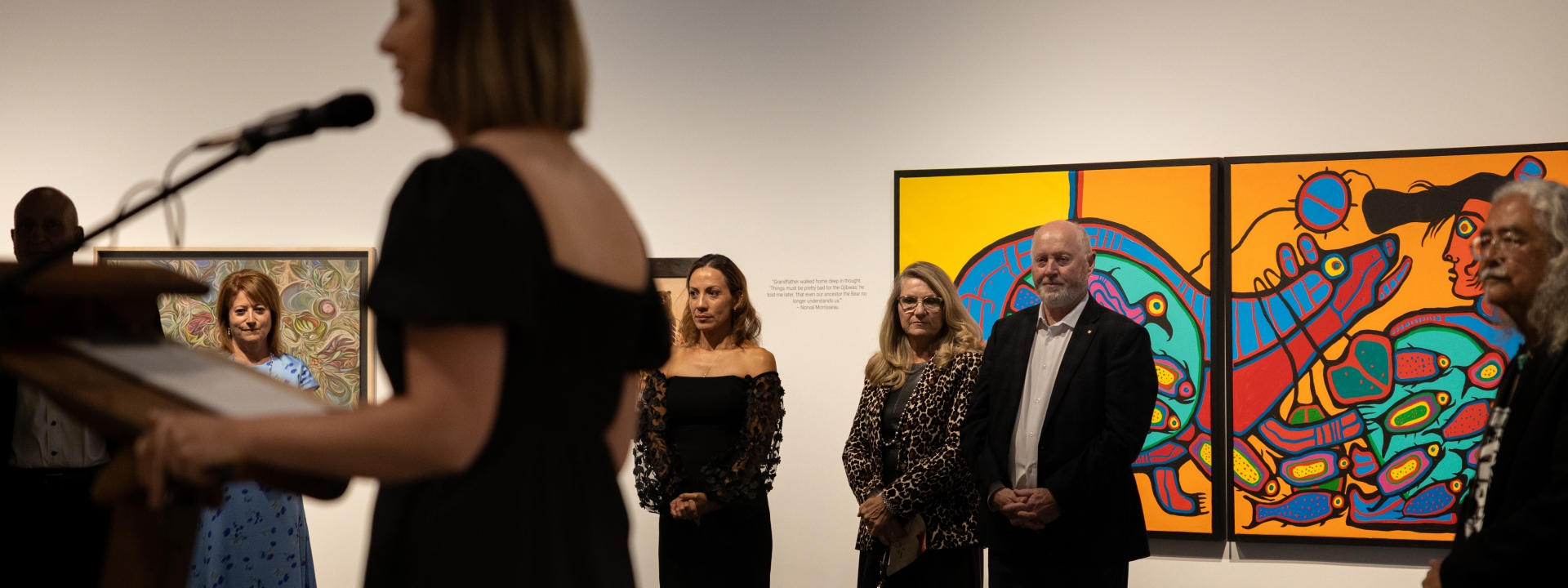 A speaker at a podium addressing attendees in an art gallery at The Whyte Museum, with guests standing in front of vibrant Indigenous artwork on the walls.