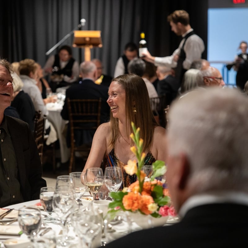 Guests seated at a dinner table at The Whyte Museum, smiling and talking while servers pour wine in the background during an evening event.