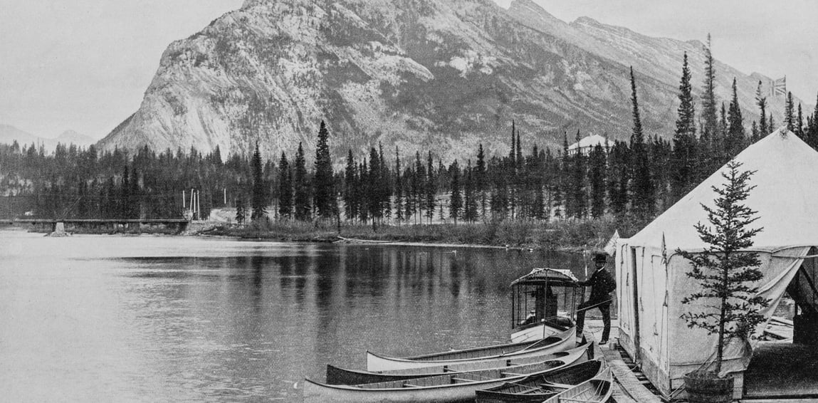 Historic photograph of canoes lined along the Bow River near Banff, with a man by a canvas tent and Mount Rundle rising in the background.