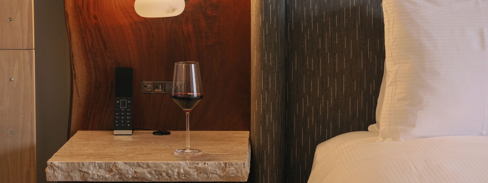 A bed from Rimrock Banff, Emblems Collection with a side stand and glass of red wine.