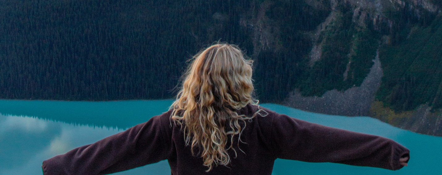 Woman sitting on a rock with arms outstretched above turquoise Lake Louise at sunrise, viewed from the Little Beehive trail.