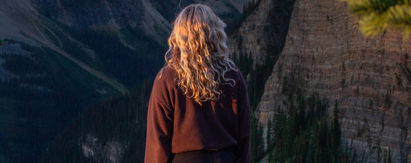 Woman standing on a rocky overlook framed by alpine trees, facing sunlit peaks along the Little Beehive trail near Lake Louise.